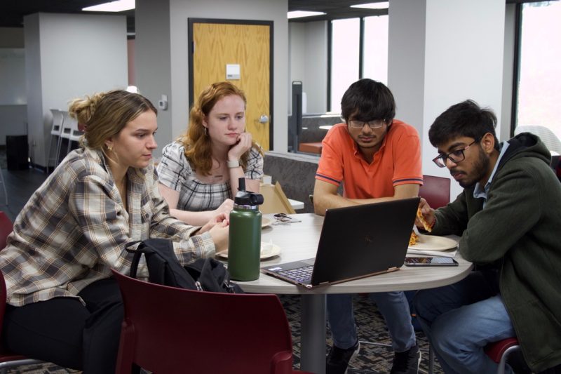 Four students seated and gathered around a laptop on a table.